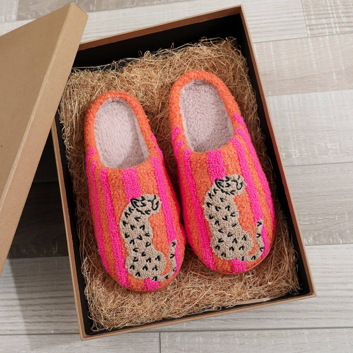 A pair of handmade knit slipper shoes in a brown gift box with natural fiber padding, set on a light wood floor. The slipper features a bright pink and orange striped knit body with a black and white spotted leopard design on the side.