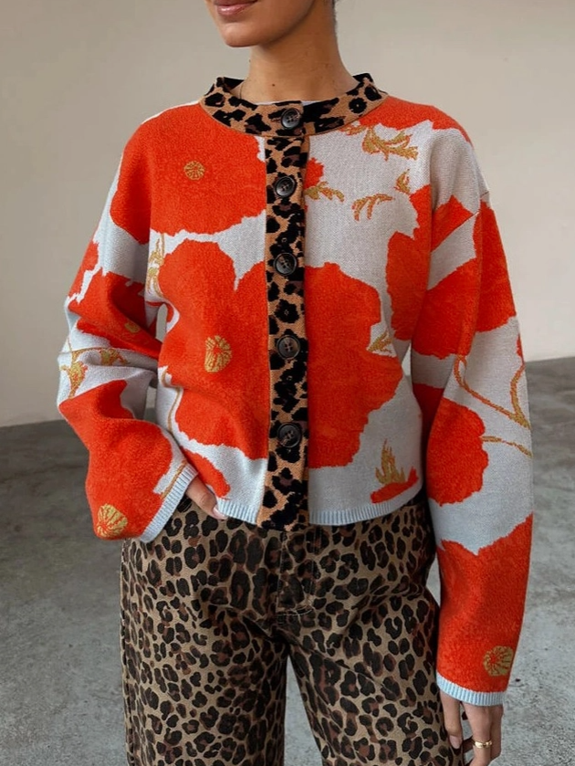 A person wearing a cropped knit cardigan with a bold orange, white, and black abstract floral pattern, leopard-print trim and button placket, paired with leopard-print pants, photographed indoors against a plain wall.