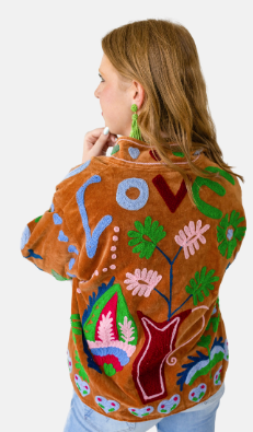 A woman shown from the back wearing a brown suede-like jacket decorated with colorful embroidered floral and abstract patterns in green, pink, blue, red, and white, against a plain light gray background.
