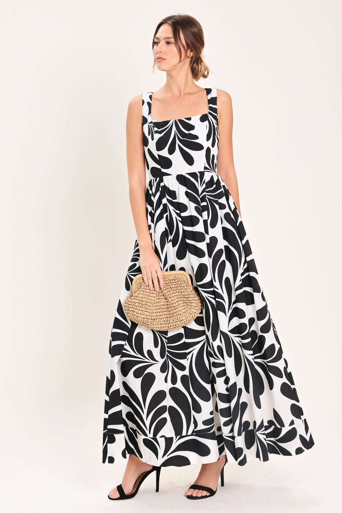 Woman wearing a black and white patterned dress holding a straw clutch against a white background