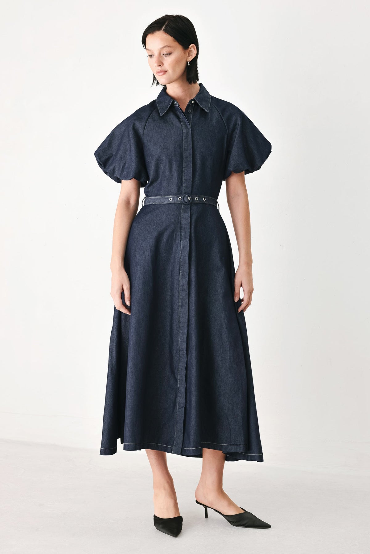 A woman modeling a dark navy blue midi shirt dress with short puff sleeves, a pointed collar, and a matching dark belt with silver studs, paired with black pointed-toe heels against a plain white studio background.