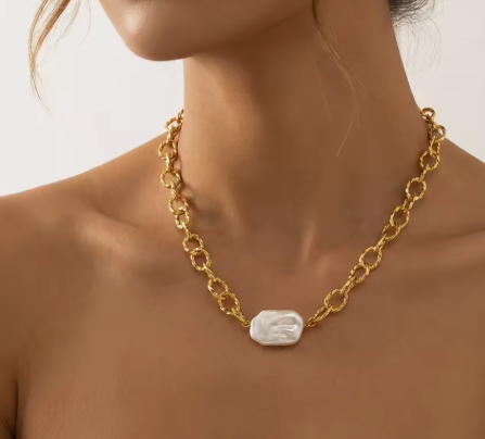 A close-up of a model wearing a gold chain necklace with a central white baroque shell pendant.
