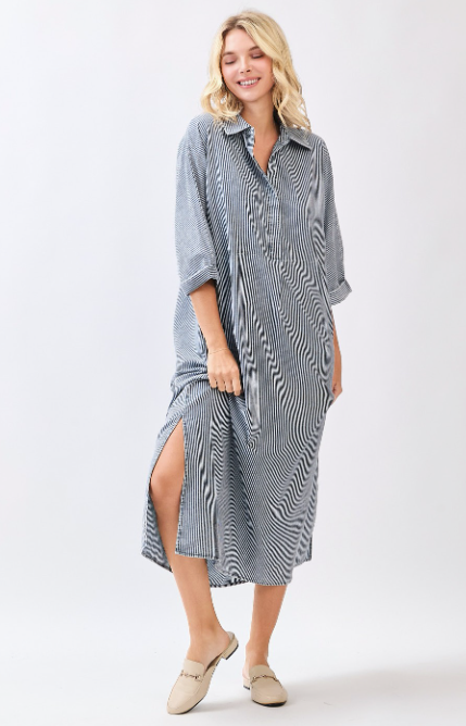 A woman modeling a loose, long-sleeve midi dress with a gray and white wavy stripe pattern, featuring a high side slit and beige slip-on mules, set against a plain light gray studio background.