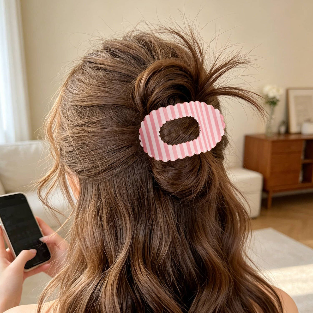 A pink and white striped hair claw clip holding a half-up hairstyle in a woman's brown hair, shown in a cozy indoor living room setting.