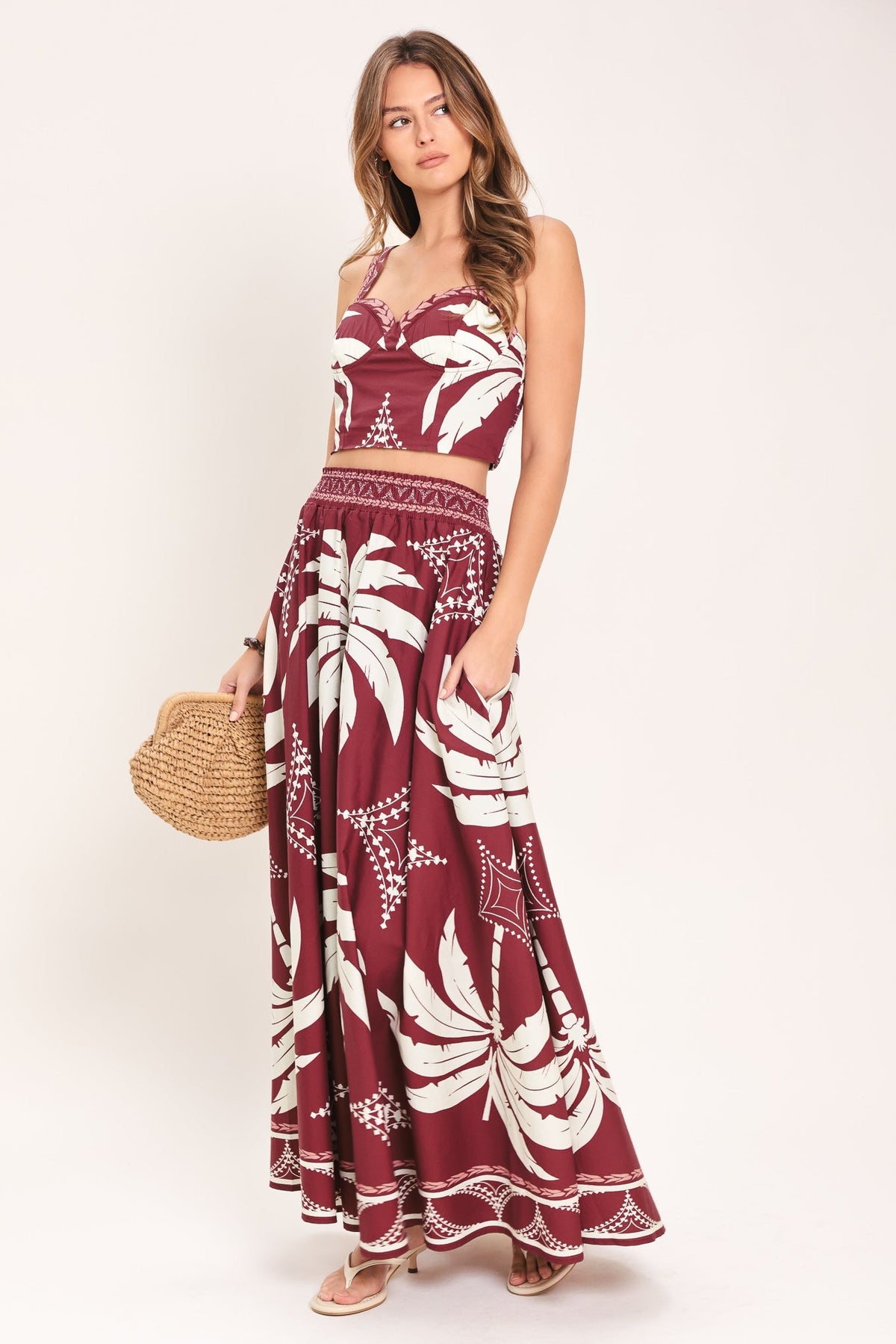 Woman wearing a red and white patterned dress holding a straw bag on a white background