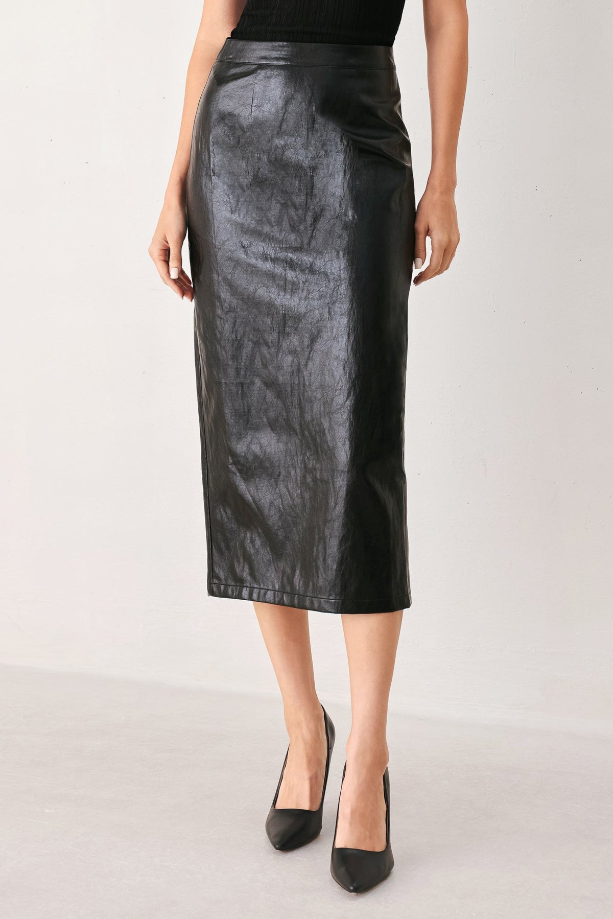 A black faux-leather midi skirt worn by a model, paired with black pointed-toe heels, shown against a plain light gray studio background.