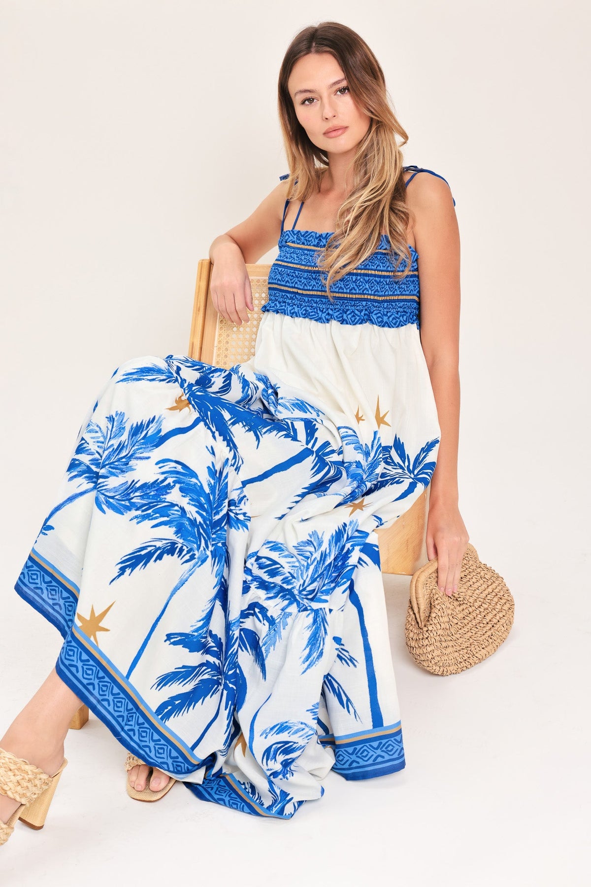 Woman wearing a blue and white dress with palm tree design, holding a straw bag on a white background