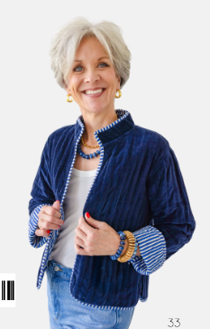 A smiling older woman with short gray hair wears a navy blue quilted jacket with striped cuffs and white trim, over a white top and light blue jeans, against a plain light background.