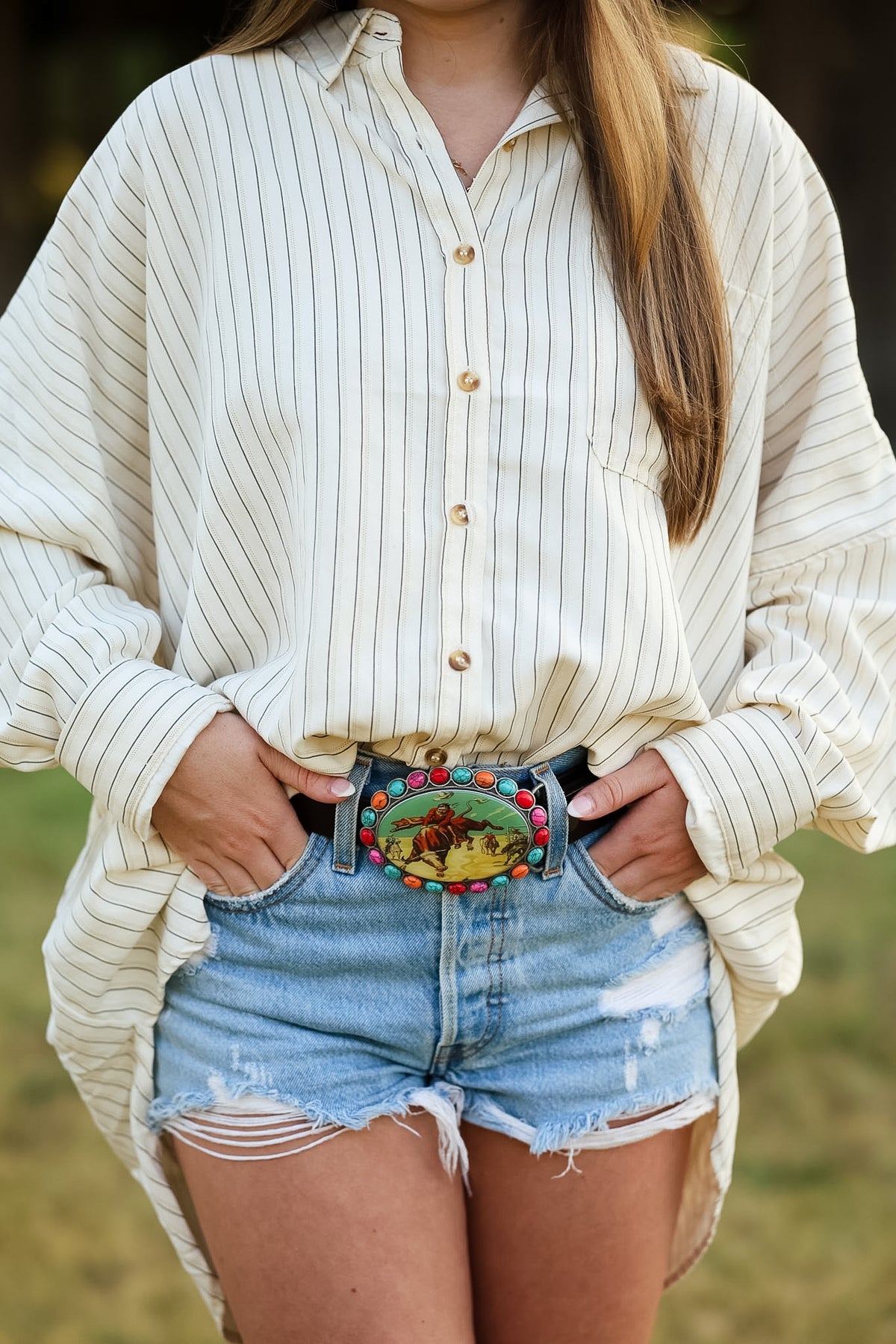 A person wearing a cream and tan vertically striped button-up shirt and light blue distressed denim shorts, featuring a decorative multicolor belt with a cowboy buckle.