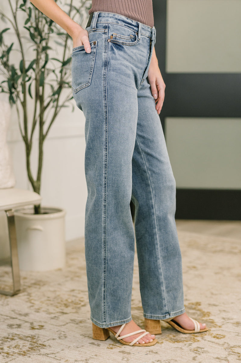 Person wearing light blue jeans standing indoors with a plant and chair in the background.