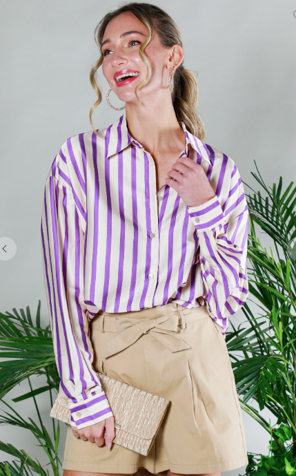Woman wearing a purple and white striped shirt with beige shorts, standing in front of green plants.