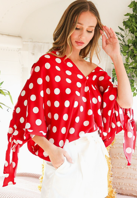 Woman wearing a red polka dot blouse with white pants, sitting on a woven chair.