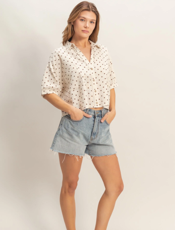 Woman wearing a white blouse with small patterns and denim shorts on a plain background