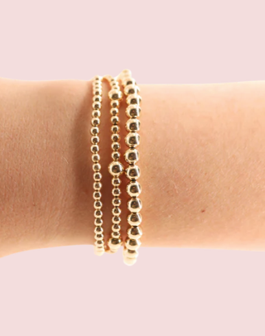 Gold beaded bracelets on a wrist against a light pink background