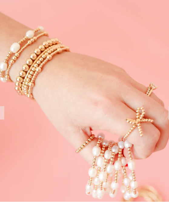 Hand wearing multiple gold and pearl bracelets on a pink background