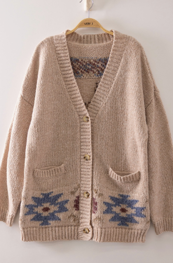 A beige knit cardigan sweater with blue and brown geometric patterns near the hem, hanging on a wooden hanger against a plain white wall.