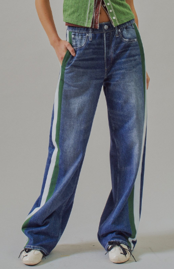 A person wearing wide-leg blue denim jeans with bold green and white side stripes, paired with a green top and white sneakers, photographed against a plain light gray studio background.