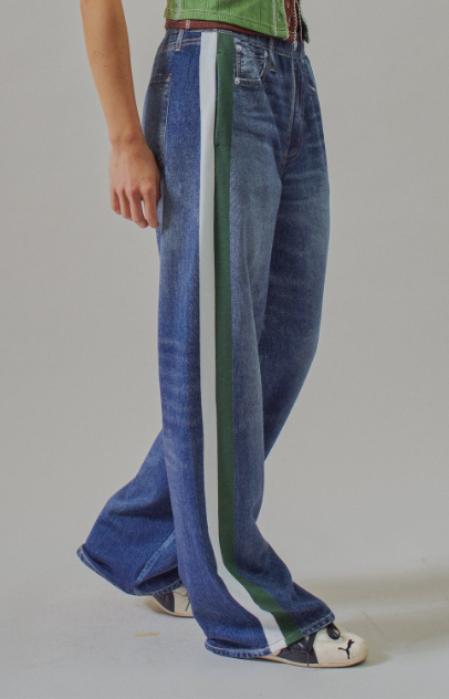 A person wearing wide-leg blue denim jeans with a bold vertical side stripe in green, white, and blue, paired with a green top and white sneakers, photographed against a plain light gray studio background.