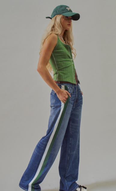 A woman modeling a green ribbed knit sleeveless top and wide-leg blue jeans with a green and white side stripe, paired with a green baseball cap, against a plain light gray studio background.