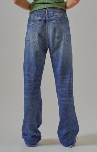 Rear view of a person wearing loose-fit blue denim jeans with a green top, standing against a plain light gray studio background.