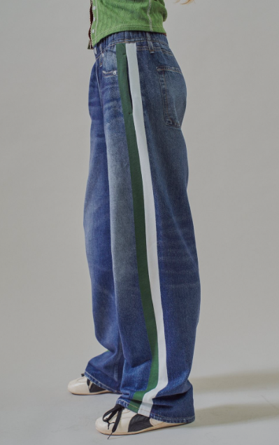 A side view of a person wearing wide-leg blue denim jeans with a vertical green and white stripe down the side, paired with a green top and black-and-white sneakers, against a plain light gray studio background.