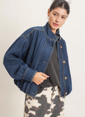 A woman wearing a cropped blue denim jacket with a high collar, button front, and voluminous sleeves, styled over a black top and a black-and-white cow-print skirt, against a plain light background.