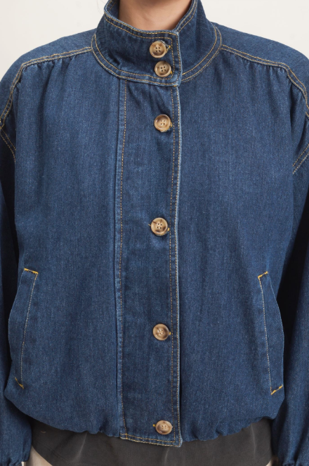A close-up of a blue denim jacket with a high stand-up collar, front button closure, and side pockets, shown on a model against a plain light background.
