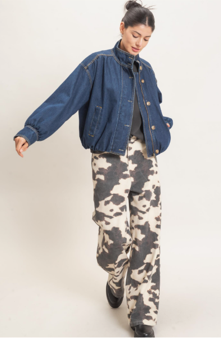 A model wearing a cropped blue denim jacket, a black top, and wide-leg cropped pants with a black and beige cow print, walking against a plain light gray studio background.