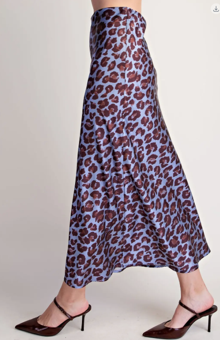 A model wearing a high-waisted, flowing midi skirt with a brown leopard print on a dusty lavender-blue background, paired with dark brown pointed-toe mule heels against a plain light gray studio background.