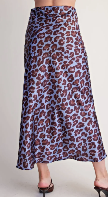 A model's lower body wearing a high-waisted, midi-length A-line skirt with a brown leopard print on a dusty lavender-blue background, paired with black strappy heels against a plain off-white studio background.