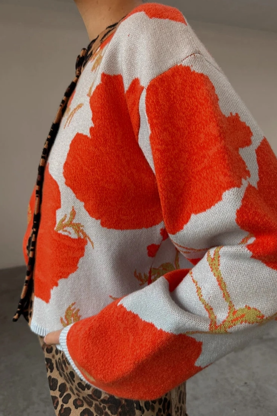 A close-up of a person wearing a fuzzy knit cardigan with a bold orange and off-white floral pattern, paired with a leopard-print bottom.