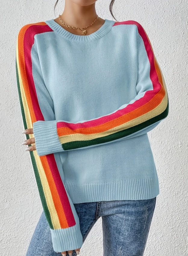A woman wearing a light blue crewneck sweater with long sleeves featuring bold rainbow stripes in red, orange, yellow, green, and pink, paired with blue jeans against a textured white wall.