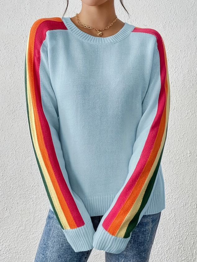 A person wearing a light blue crewneck sweater with rainbow-striped sleeves, paired with blue jeans and a gold necklace, standing against a textured off-white wall.
