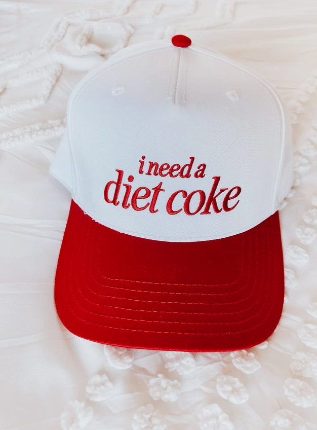A red and white trucker-style baseball cap with the phrase 'i need a diet coke' embroidered in red script across the front white panel, photographed on a white textured fabric background.