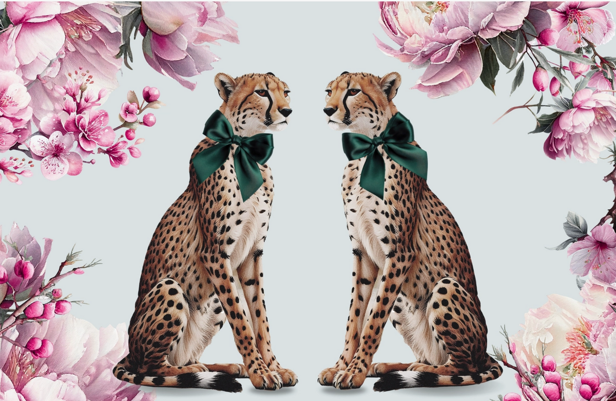 Two cheetahs sitting side by side, each wearing a dark green bow, framed by large pink flowers on a pale blue-gray background.