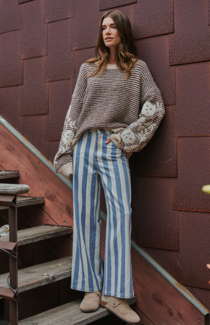 A woman stands on rustic wooden stairs against a reddish-brown textured wall, wearing a beige and brown striped knit sweater with cream knit cuffs, blue and white vertically striped wide-leg pants, and beige slip-on shoes.