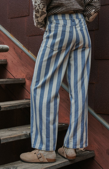 A person wearing blue and off-white vertically striped wide-leg pants, a brown patterned knit sweater, and tan Birkenstock sandals, standing on rustic wooden stairs against a red-brown wall.