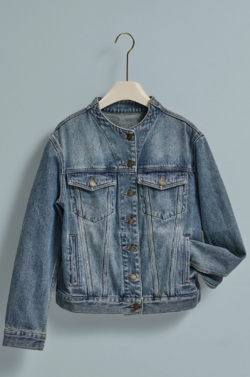 A light blue denim jacket with a raw, cropped hem and button front, hanging on a wooden hanger against a solid light blue-gray background.