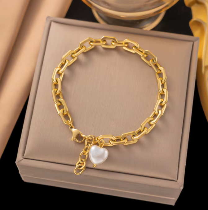 A gold chain bracelet with a dangling white heart-shaped pearl charm, displayed on a beige jewelry box.