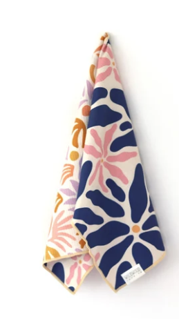 A folded cotton kitchen towel with a colorful floral and botanical pattern, featuring navy blue, pink, orange, and purple motifs on a cream background, shown against a plain white wall.