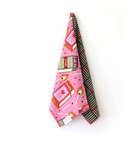 A folded pink fabric accessory, likely a scarf or bandana, featuring a Valentine's Day themed pattern with books, matches, and hearts. The reverse side shows a black and green striped pattern, displayed against a clean white background.