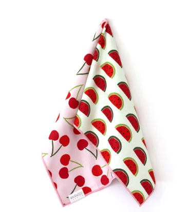 A folded cotton kitchen towel with a watermelon and cherry print, featuring a white background with red watermelon slices and pink sections with red cherries, on a white background.