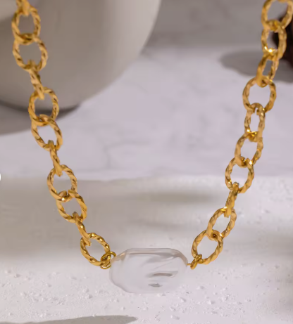 A close-up of a gold chain necklace with a clear, faceted stone pendant resting on a white marble-like surface.