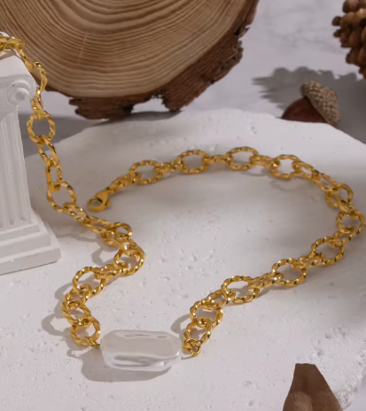 A gold chain necklace with a clear, polished teardrop-shaped stone pendant, displayed on a textured white surface with rustic wood and a white column prop in the background.