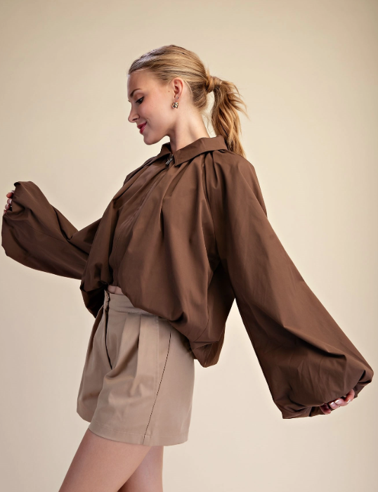 A woman wearing a brown long-sleeve blouse with dramatic balloon sleeves and beige shorts, posing against a solid beige studio background.