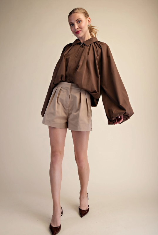 A woman modeling a brown long-sleeve blouse with dramatic balloon sleeves and beige pleated shorts against a plain beige studio background.