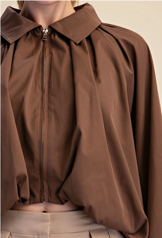 A close-up of a model wearing a brown long-sleeve blouse with a collar, front zipper, and dramatic gathered puff sleeves, paired with beige trousers against a warm beige background.