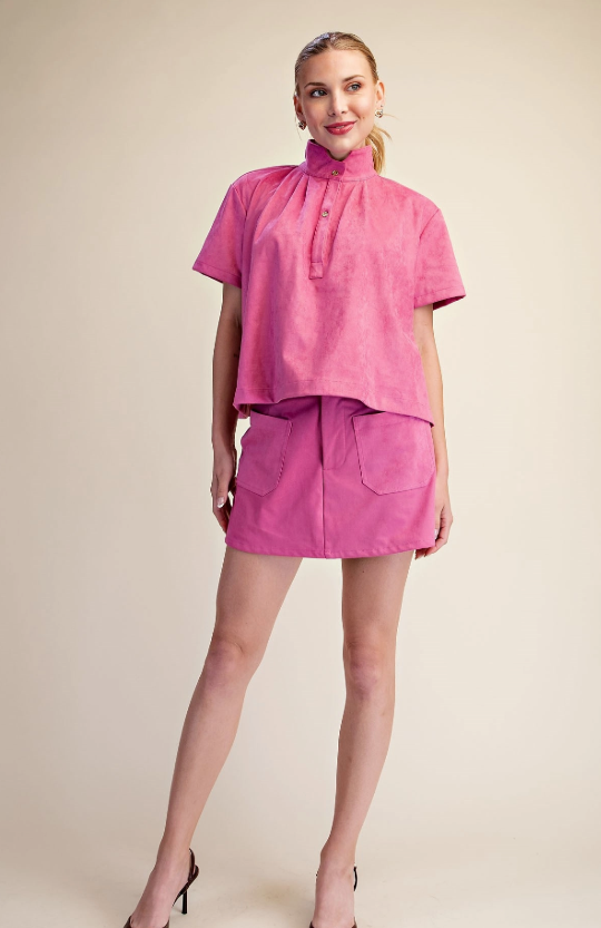 A woman modeling a bright pink two-piece outfit consisting of a short-sleeve mock-neck top and matching high-waisted mini skirt with front patch pockets, set against a solid beige studio background.