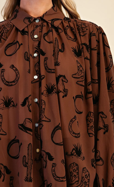 A close-up of a brown button-up blouse with a black western-themed print featuring horseshoes, cacti, cowboy boots, and hats, worn by a model.