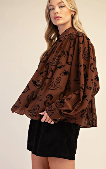 A woman wearing a brown long-sleeve blouse with a black western-themed print of boots, hats, and cacti, paired with a black skirt, against a plain beige background.